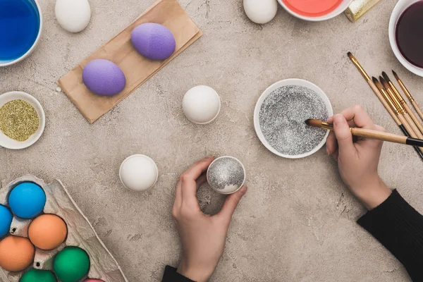 Vista cortada de mulher decorando ovos de galinha com brilho de prata na superfície de concreto cinza — Fotografia de Stock