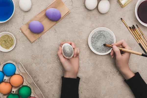 Vue recadrée d'une femme décorant des œufs de poulet avec des paillettes argentées sur une surface en béton gris — Photo de stock