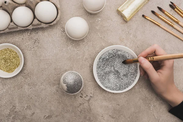 Vue recadrée d'une femme décorant un œuf de poulet avec des paillettes argentées sur une surface en béton gris — Photo de stock