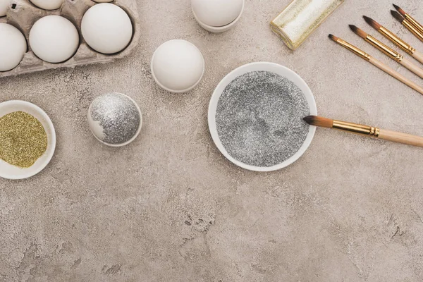 Top view of chicken eggs, silver and golden glitter with paintbrushes on grey concrete surface — Stock Photo