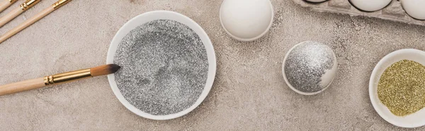 Top view of chicken eggs, silver and golden glitter with paintbrushes on grey concrete surface, panoramic shot — Stock Photo