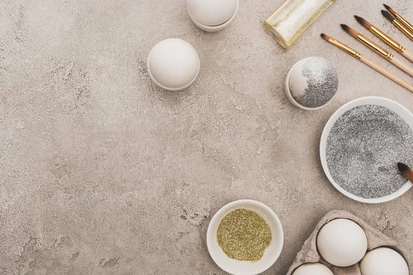 Top view of chicken eggs, silver and golden glitter with paintbrushes on grey concrete surface — Stock Photo