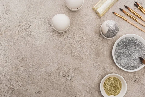 Top view of chicken eggs, silver and golden glitter with paintbrushes on grey concrete surface — Stock Photo