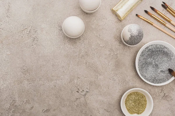 Vue de dessus des œufs de poulet, argent et paillettes dorées avec pinceaux sur la surface en béton gris — Photo de stock