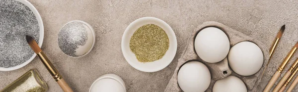 Top view of chicken eggs, silver and golden glitter with paintbrushes on grey concrete surface, panoramic shot — Stock Photo