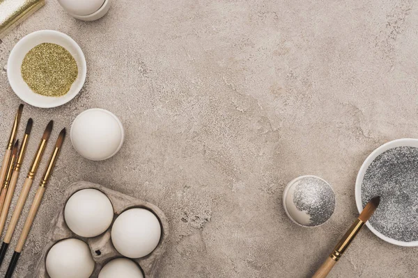 Top view of chicken eggs, silver and golden glitter with paintbrushes on grey concrete surface — Stock Photo
