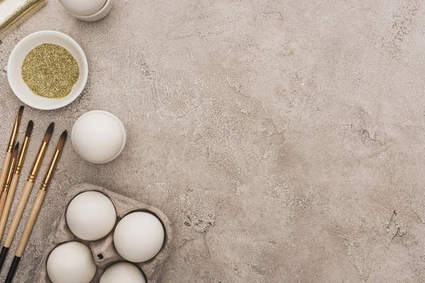 Top view of chicken eggs, golden glitter with paintbrushes on grey concrete surface — Stock Photo