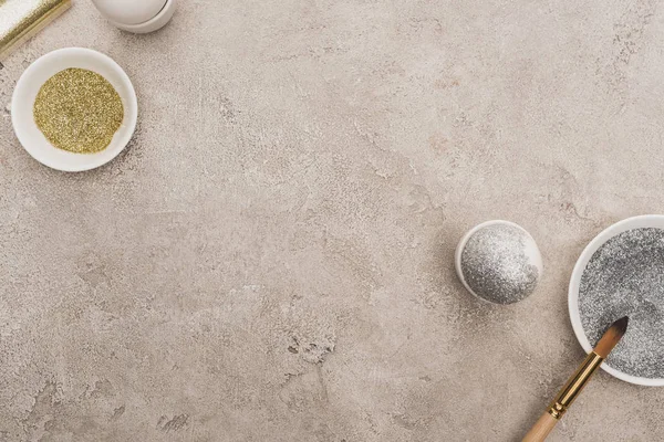 Vista superior de huevos de pollo, plata y purpurina dorada con pinceles sobre superficie de hormigón gris - foto de stock