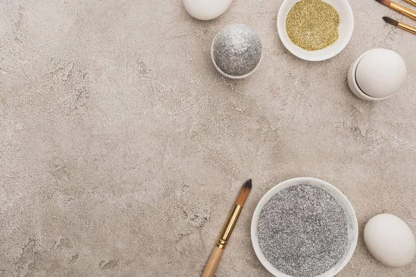 Top view of chicken eggs, silver and golden glitter with paintbrushes on grey concrete surface — Stock Photo