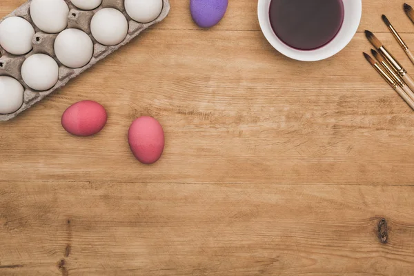 Vue du dessus de la peinture pourpre aquarelle dans un bol près des œufs de poulet peints et des pinceaux sur une table en bois — Photo de stock