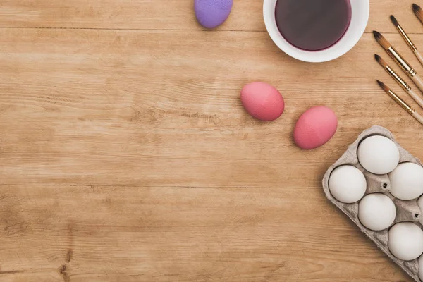 Vue du dessus de la peinture pourpre aquarelle dans un bol près des œufs de poulet et des pinceaux sur une table en bois — Photo de stock