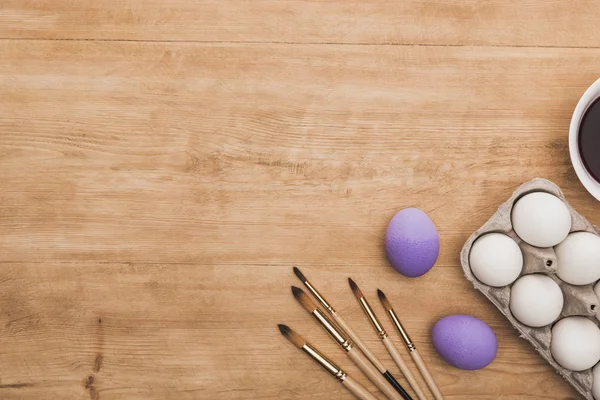 Vue du dessus de la peinture pourpre aquarelle dans un bol près des œufs de poulet et des pinceaux sur une table en bois — Photo de stock