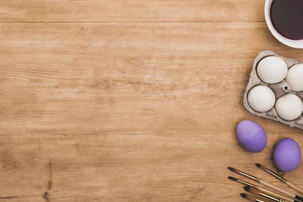 Top view of watercolor purple paint in bowl near chicken eggs and paintbrushes on wooden table — Stock Photo