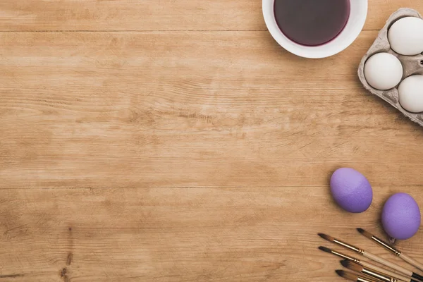 Vista dall'alto della vernice viola ad acquerello in ciotola vicino alle uova di pollo e pennelli sul tavolo di legno — Foto stock