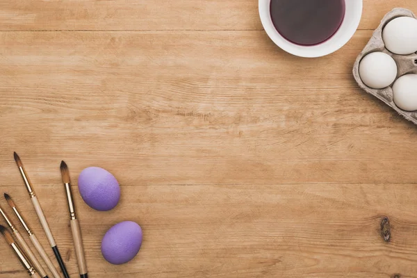 Vue du dessus de la peinture pourpre aquarelle dans un bol près des œufs de poulet et des pinceaux sur une table en bois — Photo de stock