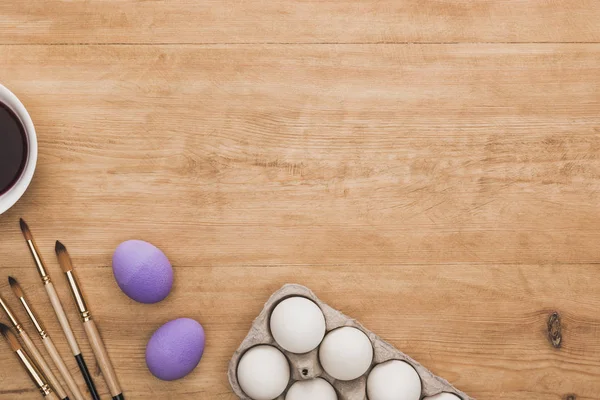 Top view of watercolor purple paint in bowl near chicken eggs and paintbrushes on wooden table — Stock Photo