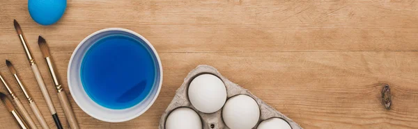 Вид зверху акварельної синьої фарби в мисці біля курячих яєць і пензлів на дерев'яному столі, панорамний знімок — стокове фото