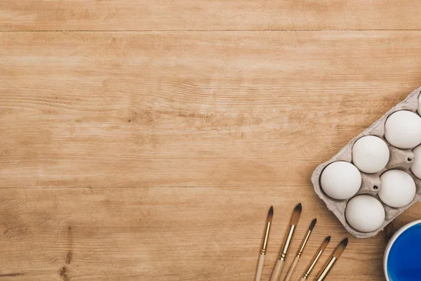 Top view of watercolor blue paint in bowl near chicken eggs and paintbrushes on wooden table — Stock Photo