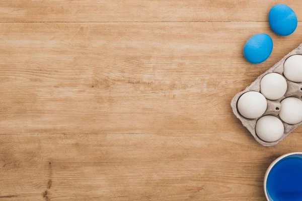 Vue du dessus de la peinture bleue aquarelle dans un bol près des œufs de poulet et des pinceaux sur une table en bois — Photo de stock