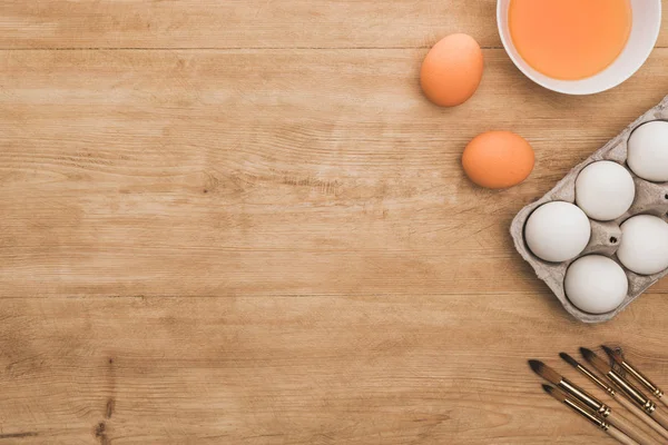 Вид сверху на акварельную оранжевую краску в миске возле куриных яиц и кистей на деревянном столе — стоковое фото
