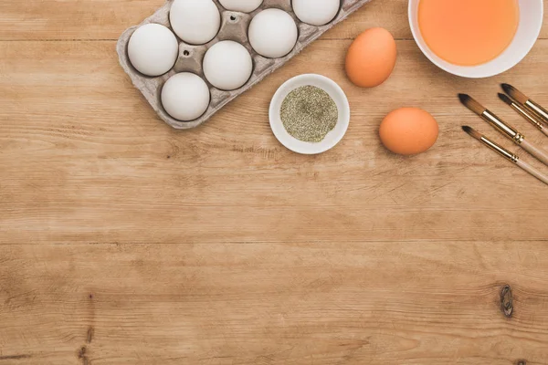 Вид сверху на оранжевую акварельную краску и золотой блеск в чашах возле пасхальных яиц и кистей на деревянном столе — стоковое фото