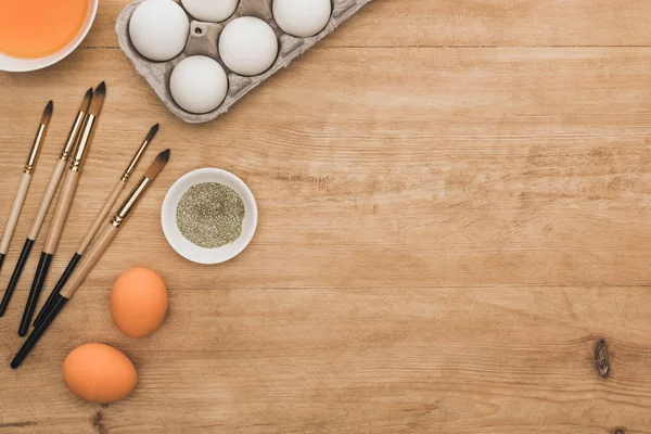 Vue du dessus de peinture à l'aquarelle orange et paillettes dorées dans des bols près des oeufs de Pâques et pinceaux sur table en bois — Photo de stock