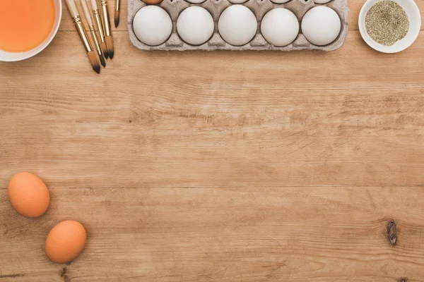 Vista dall'alto di vernice arancione ad acquerello e brillantini dorati in ciotole vicino alle uova di Pasqua e pennelli sul tavolo di legno — Foto stock