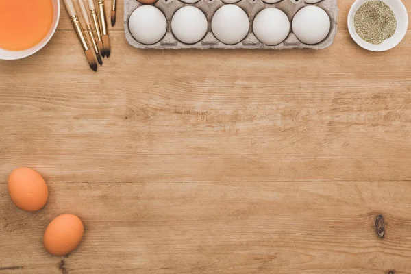 Vista superior de pintura de acuarela naranja y brillo dorado en cuencos cerca de huevos de Pascua y pinceles en mesa de madera - foto de stock