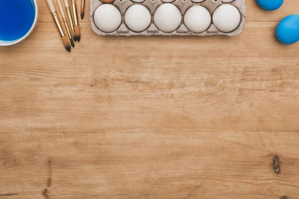 Top view of blue watercolor paint in bowl near chicken eggs and paintbrushes on wooden table — Stock Photo