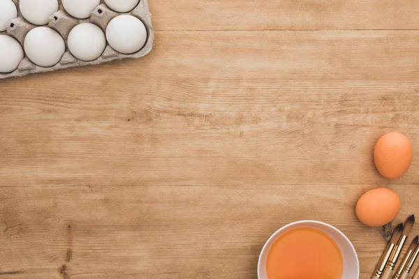 Draufsicht auf orangefarbene Aquarellfarbe in Schüssel in der Nähe von Hühnereiern und Pinseln auf Holztisch — Stockfoto