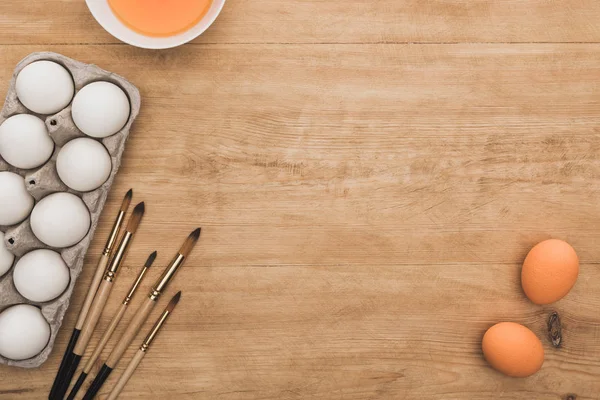Вид зверху апельсинової акварельної фарби в мисці біля курячих яєць і пензлів на дерев'яному столі — стокове фото
