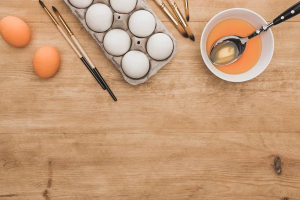 Вид апельсиновой акварельной краски в миске возле куриных яиц и кистей на деревянном столе — стоковое фото
