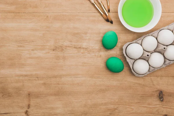 Top view of green watercolor paint in bowl near chicken eggs and paintbrushes on wooden table — Stock Photo