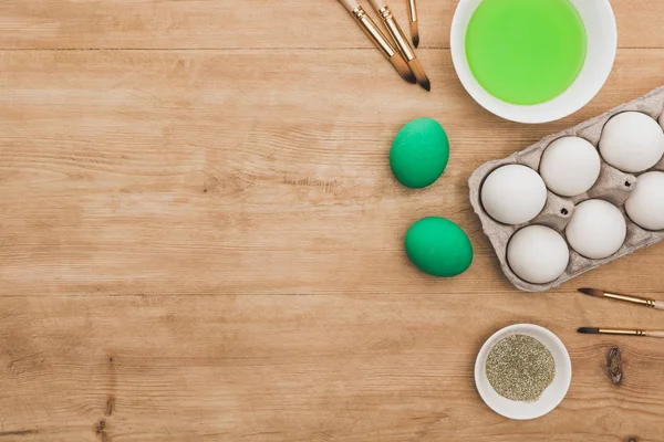Vista dall'alto di vernice verde ad acquerello e brillantini in ciotole vicino alle uova di Pasqua e pennelli sul tavolo di legno — Foto stock