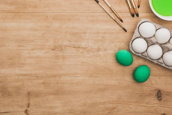 Top view of green watercolor paint in bowl near chicken eggs and paintbrushes on wooden table — Stock Photo