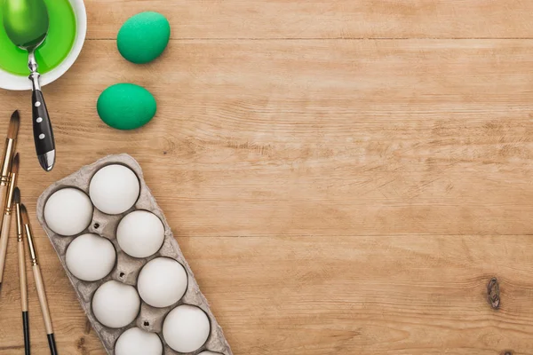 Top view of green watercolor paint in bowl near chicken eggs and paintbrushes on wooden table — Stock Photo