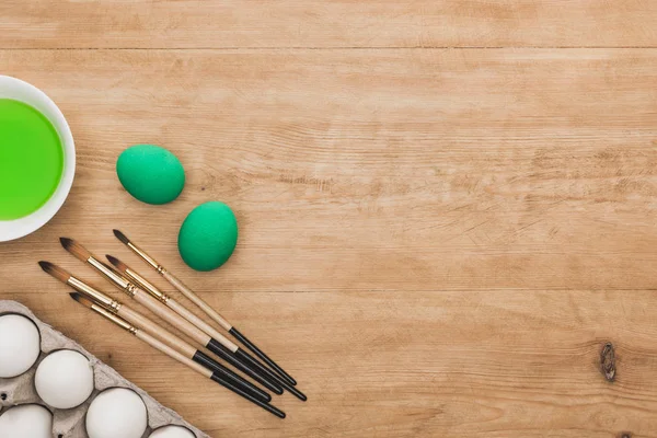 Вид зеленой акварельной краски в миске рядом с куриными яйцами и кисточками на деревянном столе — стоковое фото