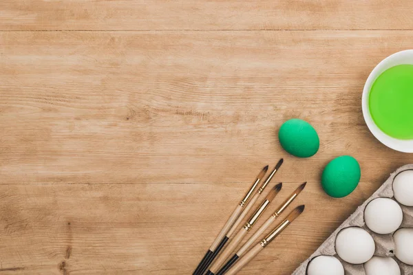 Vue du dessus de la peinture à l'aquarelle verte dans un bol près des œufs de poulet et des pinceaux sur une table en bois — Photo de stock