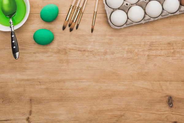 Vista dall'alto della vernice verde ad acquerello in ciotola vicino alle uova di pollo e pennelli sul tavolo di legno — Foto stock