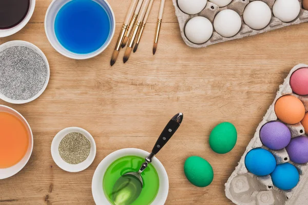 Top view of watercolor paints and glitter in bowls near Easter eggs and paintbrushes on wooden table — Stock Photo