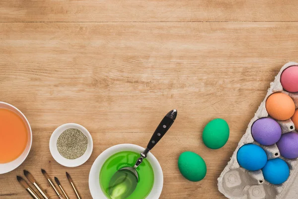 Верхний вид акварельных красок и блеска в чашах возле пасхальных яиц и кистей на деревянном столе — стоковое фото