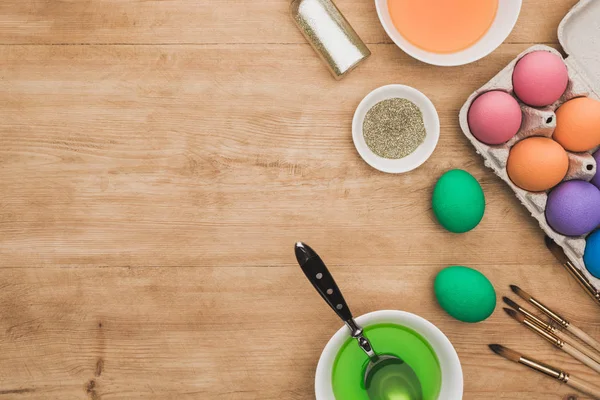 Vue de dessus des peintures aquarelles et des paillettes dans des bols près des oeufs de Pâques et des pinceaux sur la table en bois — Photo de stock