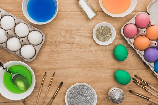 Top view of watercolor paints and glitter in bowls near Easter eggs and paintbrushes on wooden table — Stock Photo