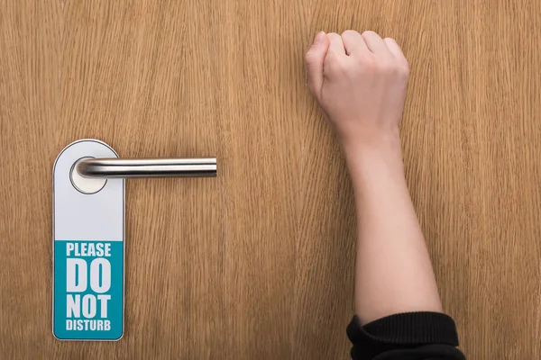 Cropped view of woman knocking at door with please do not disturb sign — стоковое фото