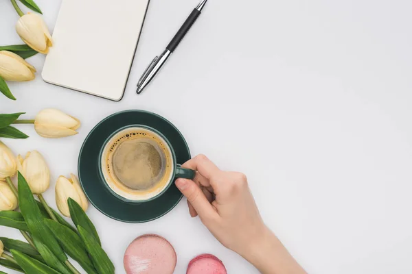 Vista recortada de la mujer sosteniendo taza de café cerca de tulipanes, cuaderno en blanco y deliciosos macarons aislados en blanco - foto de stock