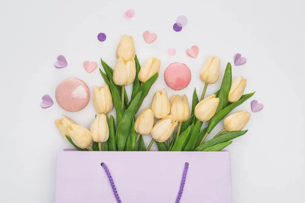 Vista superior de tulipas, macarons rosa e corações violetas em saco de compras isolado em branco — Fotografia de Stock