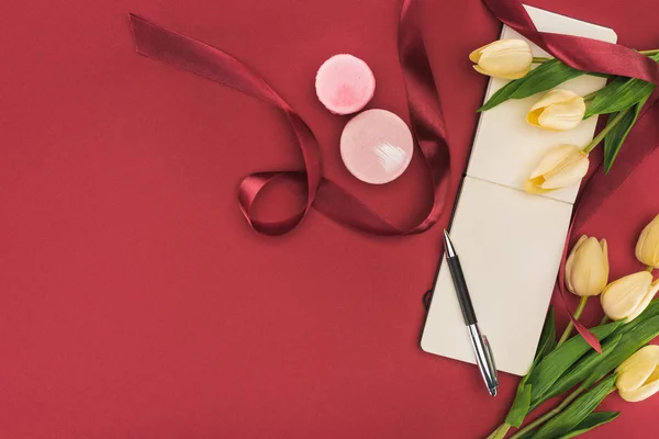 Top view of tulips with silk ribbon, macarons and empty notebook with pen isolated on red — Stock Photo