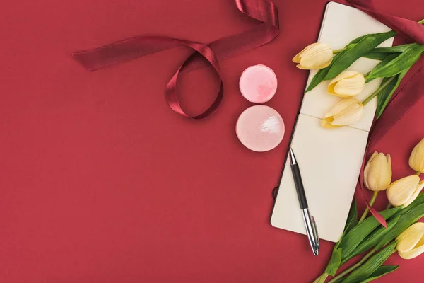 Top view of tulips with silk ribbon, macarons and empty notebook with pen isolated on red — Stock Photo