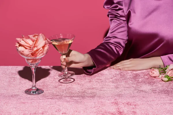 Cropped view of woman holding glass of rose wine near glass with rose petals on velour cloth isolated on pink, girlish concept — Stock Photo