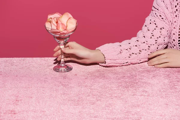 Vista recortada de la mujer sosteniendo vidrio con pétalos de rosa en tela de terciopelo aislado en rosa, concepto femenino - foto de stock